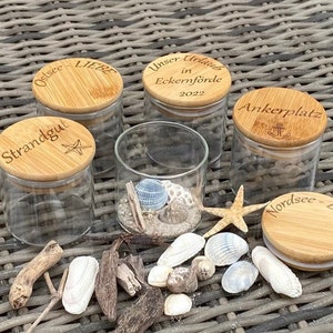 Könnte beinhalten: Klare Glasbehälter mit Bambusdeckeln, einige gefüllt mit Sand und Muscheln. Die Deckel sind mit Sätzen wie "Ostsee Liebe" und "Strandgut" graviert. Ein Seestern und verschiedene Muscheln sind um die Gläser angeordnet.