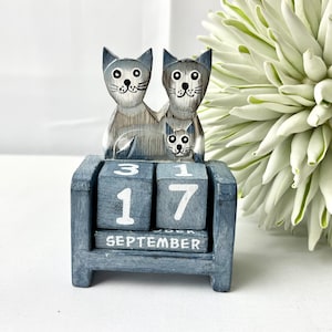 Pode incluir: Calendário perpétuo de madeira cinza com design de família de gatos. O calendário exibe "17 SETEMBRO" em seus blocos rotativos. As figuras de gatos são esculpidas à mão com detalhes pintados. É um item decorativo.