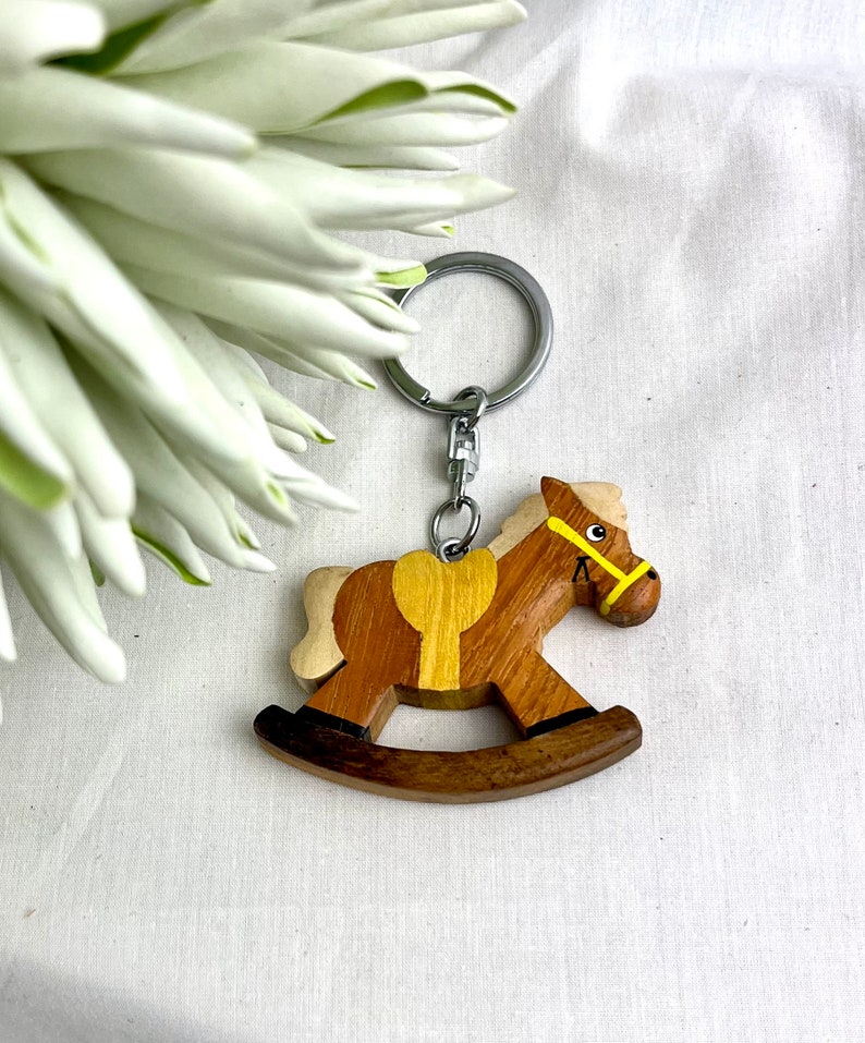 May include: A wooden rocking horse keychain with a silver keyring. The horse is brown with a yellow saddle and bridle. The keychain is a charming accessory, perfect for anyone who loves horses or vintage toys.