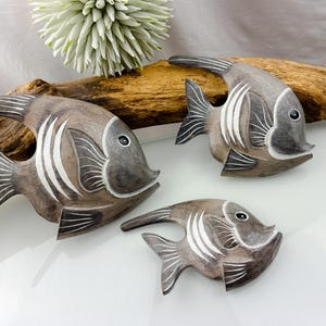 May include: Three wooden fish sculptures in varying sizes, each with a grey and brown finish and white accent lines. The fish are arranged on a white surface with a piece of driftwood and a white flower in the background. Decorative home decor.