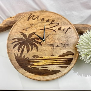 May include: Round wooden wall clock with a beach scene design. The clock features a palm tree, seagulls, and the text "Meer Zeit". The clock hands are black.
