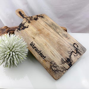 May include: Wooden cutting board with a handle, featuring a natural wood grain and dark, tree-like patterns. The board has the text "Wunschtext..." on it. A white and green decorative ball and a piece of driftwood are also in the image.