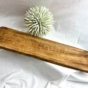 May include: A rectangular wooden serving tray with a natural wood grain finish. The tray has a slightly angled shape and a recessed area along the edges. A decorative white and green spiky ball is in the background.