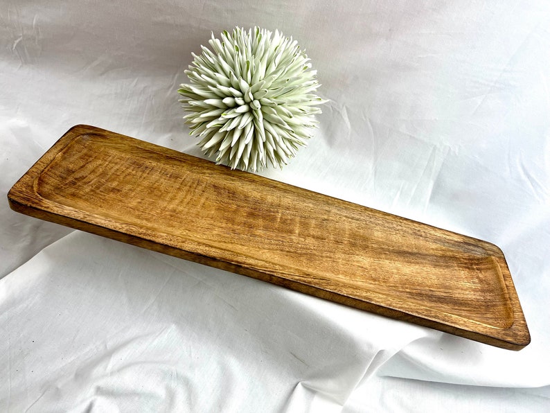 May include: A rectangular wooden serving tray with a natural wood grain finish. The tray has a slightly angled shape and a recessed area along the edges. A decorative white and green spiky ball is in the background.