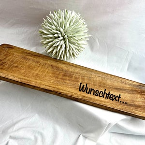 May include: A rectangular wooden serving tray with a natural wood grain finish. The tray has a slightly raised edge and the text "Wunschtext..." is etched on the surface. A decorative white and green floral accent is in the background.