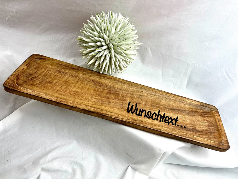 May include: A rectangular wooden serving tray with a natural wood grain finish. The tray has a slightly raised edge and the text "Wunschtext..." is etched on the surface. A decorative white and green floral accent is in the background.
