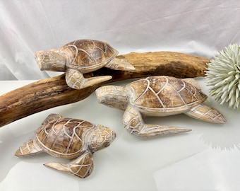 May include: Three wooden turtle figurines in varying sizes, with a light brown and cream color scheme. The turtles are positioned on a piece of driftwood, with a white background. The turtles have detailed carvings.