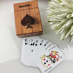 May include: A wooden playing card box with a spade design and the text "Wunschtext...". A deck of playing cards is fanned out next to the box, with the Joker card visible. The cards have black spade symbols and red and black suit symbols.