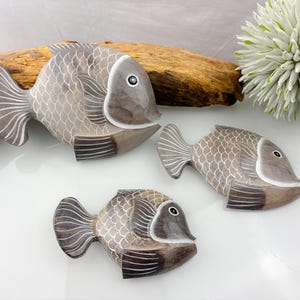 May include: Three wooden fish sculptures in shades of grey and brown, with white accents. The fish have detailed scale patterns and fins. They are arranged on a white surface with a piece of driftwood and a white flower in the background.