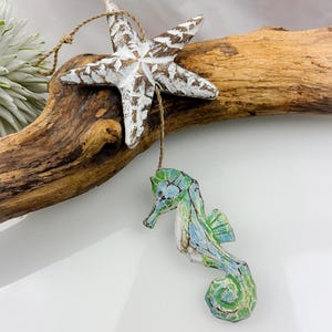 Peut inclure: Ornement en forme d'étoile de mer en bois rustique, peint en blanc, et ornement en forme d'hippocampe marron avec des détails bleus et verts, suspendus à du bois flotté. L'hippocampe est finement détaillé.