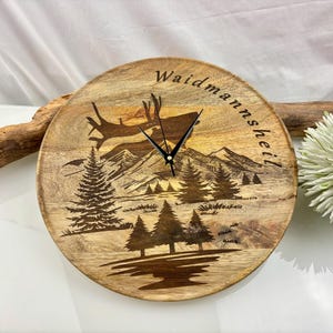 May include: A round wooden clock with a mountain and deer scene. The clock face features a detailed engraving of a forest, mountains, and a deer leaping across the sky. The clock hands are black, and the text "Waidmannsheil" is arched across the top.