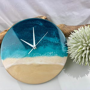 Puede incluir: Reloj de pared redondo con diseño de playa. La esfera del reloj presenta un degradado de colores, incluyendo azul oscuro, turquesa, blanco y beige, que simulan el océano y la arena. Las manecillas blancas indican la hora. Hay elementos decorativos en el fondo.