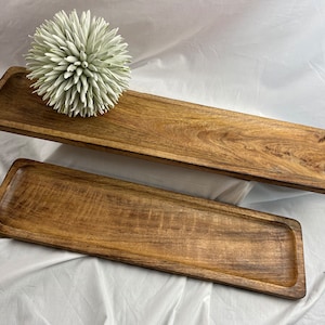 Peut inclure: Deux plateaux de service rectangulaires en bois avec une finition naturelle veinée. Les plateaux sont de tailles différentes et ont un bord légèrement surélevé. Un élément floral décoratif blanc et vert est posé sur un plateau.