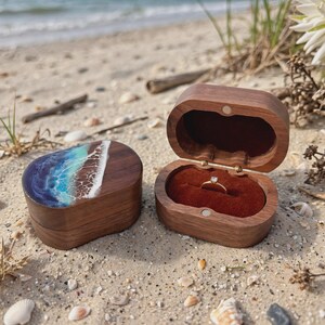 May include: Two oval wooden ring boxes made of dark walnut. One box is closed, featuring a decorative resin inlay on the lid that mimics ocean waves with blue, turquoise, and white foam. The other box is open, revealing a dark red velvet interior.