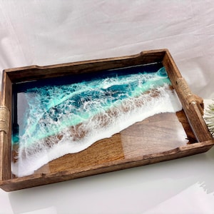 Pode incluir: Uma bandeja de servir retangular de madeira com uma incrustação de resina que imita uma costa oceânica. O design inclui tons de água azul profundo e turquesa que transitam para ondas de espuma branca, sobre uma base de madeira com alças de corda.