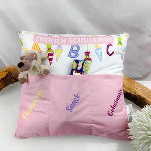 May include: A white pillow with a pink pocket, embroidered with the words "Glücksbringer", "Snack", and "Geheimnis". The pillow has a colourful design with the text "Endlich Schulkind!" and ABC letters. A stuffed animal and a snack are in the pockets.