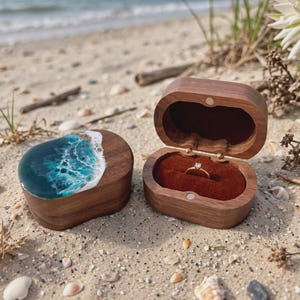 May include: Two oval wooden ring boxes with a dark brown grain. One box is closed, featuring a blue and white resin ocean wave design on the lid. The other box is open, revealing a deep red velvet interior holding a gold ring with a small stone.