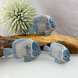 Peut inclure: Trois figurines décoratives de poissons bleus et gris avec des motifs d'écailles détaillés. Les poissons sont disposés sur un morceau de bois flotté, avec un accent floral blanc et vert en arrière-plan. Les poissons ont un design peint.