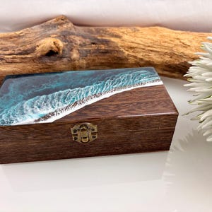 Könnte beinhalten: Eine rechteckige Holzschatulle mit einem Harzdeckel, der ein blau-weißes Meereswellen-Design aufweist. Die Schatulle hat eine dunkelbraune Holzmaserung und einen kleinen messingfarbenen Metallverschluss an der Vorderseite zum Schließen.