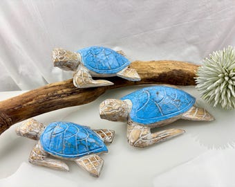 May include: Three wooden turtle figurines with blue shells and tan bodies are arranged on a piece of driftwood. The turtles have a distressed, carved appearance. A white flower is in the background.