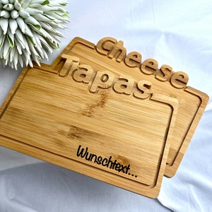 Könnte beinhalten: Zwei rechteckige Servierbretter aus Bambus. Auf dem oberen Brett sind die Worte "Cheese Tapas" eingraviert, und auf dem unteren Brett ist der Text "Wunschtext..." eingraviert. Die Bretter sind hellbraun und haben einen vertieften Rand.