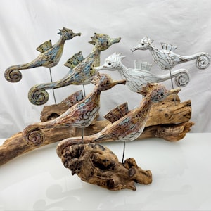 May include: Hand-carved seahorse sculptures in various colors, including blue, white, and brown, are mounted on driftwood. The seahorses have detailed fins and tails, and are displayed on a white surface against a white backdrop. The sculptures are a unique home decor item.