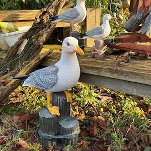 May include: Three decorative seagull statues perched on wooden posts. The seagulls have white bodies, gray wings, and yellow beaks and feet. The posts are painted to look like weathered wood, and the scene is set outdoors.