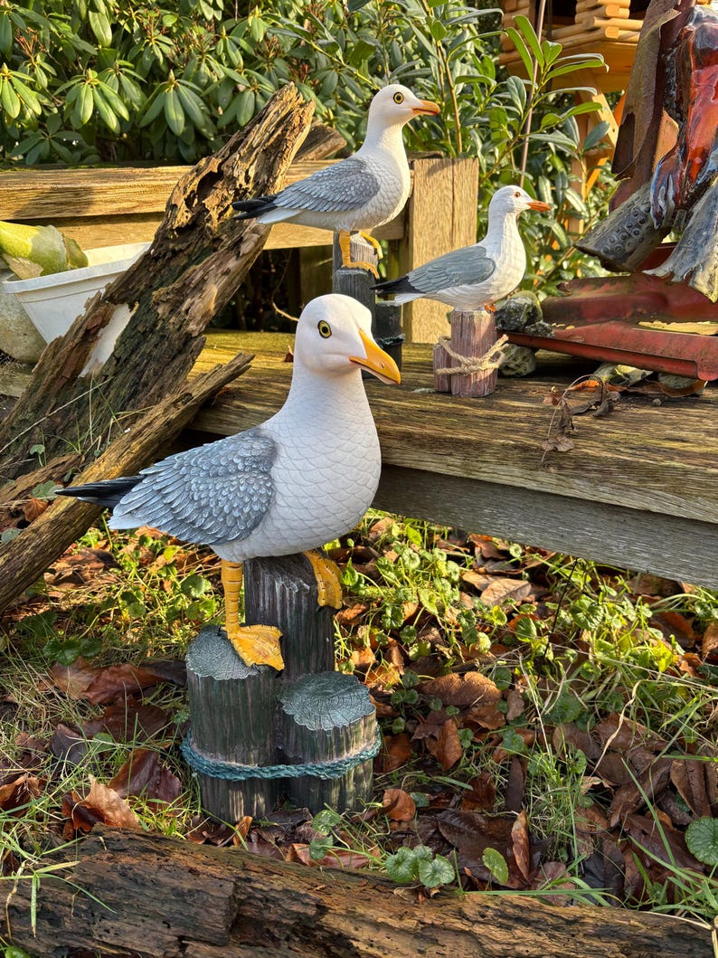 May include: Three decorative seagull statues perched on wooden posts. The seagulls have white bodies, gray wings, and yellow beaks and feet. The posts are painted to look like weathered wood, and the scene is set outdoors.