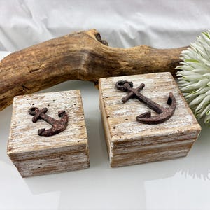 May include: Two small, rustic wooden boxes with distressed white paint and a rusty metal anchor on top. The boxes are rectangular and have a nautical theme. A piece of driftwood and a white flower are in the background.