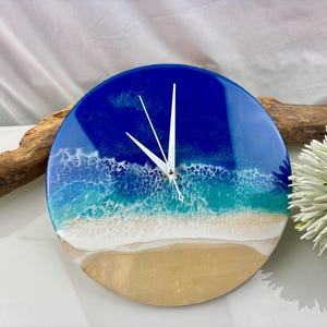 Puede incluir: Reloj de pared redondo con diseño de playa. La esfera del reloj presenta un océano azul, olas blancas y una playa de arena. Las manecillas blancas indican la hora. Es un artículo decorativo.