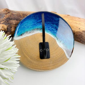 Può includere: Gancio da parete rotondo in legno con scena di spiaggia, con onde blu e bianche in resina. Il gancio è in metallo nero e fissato al legno con una piastra in metallo nero. Il legno è di colore marrone chiaro naturale.