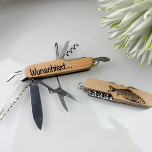 Könnte beinhalten: Zwei Schweizer Taschenmesser aus Holz mit verschiedenen Werkzeugen. Auf einem Messer steht "Wunschtext..." auf dem Griff. Das andere Messer hat ein Fischdesign. Die Messer liegen auf einer weißen Oberfläche.