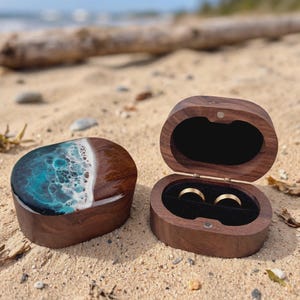 Könnte beinhalten: Zwei ovale Ringkästchen aus dunklem Walnussholz. Eine Schatulle ist geschlossen und zeigt auf dem Deckel eine glänzende Harzeinlage, die eine türkisfarbene Meereswelle mit weißem Gischt-Effekt nachahmt. Die andere ist offen und zeigt ein schwarzes Samtfutter.
