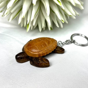 Puede incluir: Un llavero de tortuga de madera con un caparazón tallado detallado y aletas marrones oscuras. El llavero tiene un anillo de metal plateado. El fondo incluye una flor blanca y tela blanca.