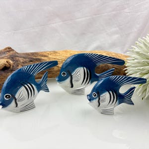 Könnte beinhalten: Drei handgeschnitzte Holzfischskulpturen. Jeder Fisch hat einen blauen Körper mit weißen und schwarzen Details, einschließlich Streifen und Flossen. Die Fische sind auf einer weißen Oberfläche angeordnet, mit Treibholz und einer weißen Blume im Hintergrund.