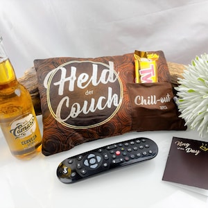 May include: A brown pillow with the text "Held der Couch" and a pocket labeled "Chill-out area" holds a candy bar. A bottle of beer, a TV remote, and a card that says "King of the Day" are also present.