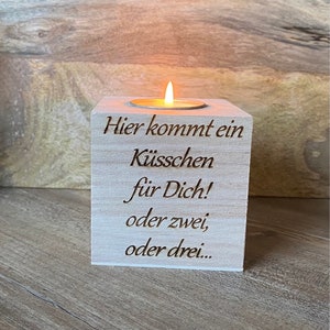 Op de afbeelding: Een houten kandelaar met een brandende waxinelichtje. De houder heeft gegraveerde tekst: "Hier kommt ein Küsschen für Dich! oder zwei, oder drei..." in een scriptlettertype. De kaarsvlam gloeit helder tegen het lichte hout.