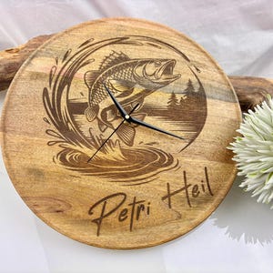 May include: A round wooden clock with a laser-etched design of a fish jumping out of water. The clock has black hands and the words "Petri Heil" are etched on the bottom. The clock is approximately 10 inches in diameter.