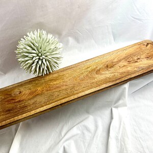 May include: A long, rectangular wooden serving tray with a natural wood grain finish. The tray has a slightly raised edge and a recessed area at each end. A decorative, spiky, light green ball is next to the tray. This tray is suitable for serving food or displaying items.