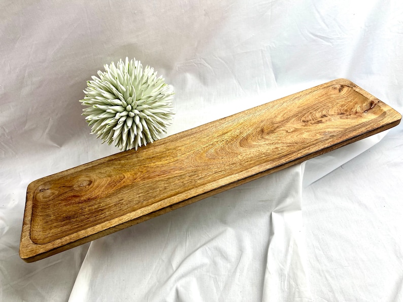 May include: A long, rectangular wooden serving tray with a natural wood grain finish. The tray has a slightly raised edge and a recessed area at each end. A decorative, spiky, light green ball is next to the tray. This tray is suitable for serving food or displaying items.