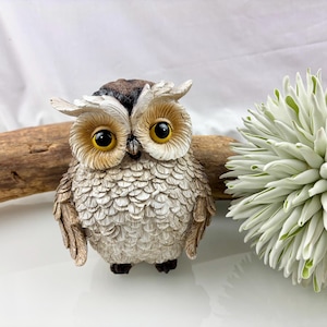 May include: A detailed figurine of an owl with large yellow eyes, brown and beige feathers, and black feet. The owl is perched on a white surface, with a piece of driftwood behind it and a white and green decorative plant to the right.