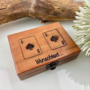May include: A wooden box with two engraved playing card aces of spades. The box is brown with a black clasp. The text "Wunschtext..." is printed on the box. The background includes a wooden branch and white flowers.
