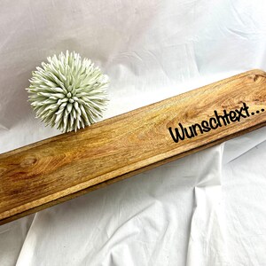May include: A rectangular wooden serving tray with a carved edge and the text "Wunschtext..." in black. The tray is made of light brown wood and has a decorative, light green, spiky ball next to it.
