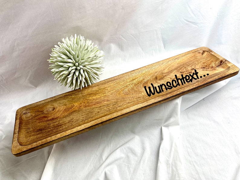 May include: A rectangular wooden serving tray with a carved edge and the text "Wunschtext..." in black. The tray is made of light brown wood and has a decorative, light green, spiky ball next to it.