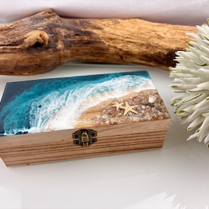 May include: A rectangular wooden box featuring a resin lid decorated with a beach scene. The design includes deep blue ocean water, white foam waves, a sandy shore, small pebbles, and two light-colored starfish. A metal latch secures the front.