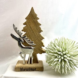 May include: A silver-colored reindeer figurine on a wooden base with the text "Wunschtext...". A wooden tree and a white and green decorative ball are behind the reindeer. The background is white.