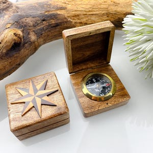 Peut inclure: Boussole en bois dans une boîte avec une lunette dorée. La boîte est ouverte, révélant le cadran de la boussole. Une deuxième boîte en bois avec un motif d'étoile est fermée. C'est un outil de navigation.