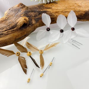 Può includere: Un assortimento di accessori per capelli con piume. Gli accessori presentano piume bianche e marroni, perline di legno e dettagli in metallo argentato. Alcuni hanno una perla rosso scuro al centro. Gli articoli sono disposti su una superficie bianca con un pezzo di legno.
