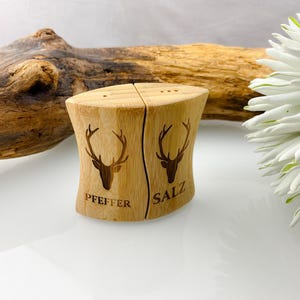 Puede incluir: Un par de saleros y pimenteros de madera. Cada salero y pimentero está hecho de madera de color claro y presenta una silueta de cabeza de ciervo. La palabra "Pfeffer" está en un salero y "Salz" en el otro.