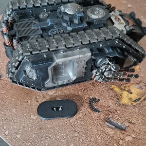 Könnte beinhalten: Ein detailliertes, dunkelgraues Modell eines Militärpanzers mit komplizierten Ketten- und Turmdetails. Das Modell enthält mehrere Kanonen und ein kleines schwarzes Teil mit der Nummer "14". Der Panzer steht auf einer strukturierten, braunen Oberfläche.